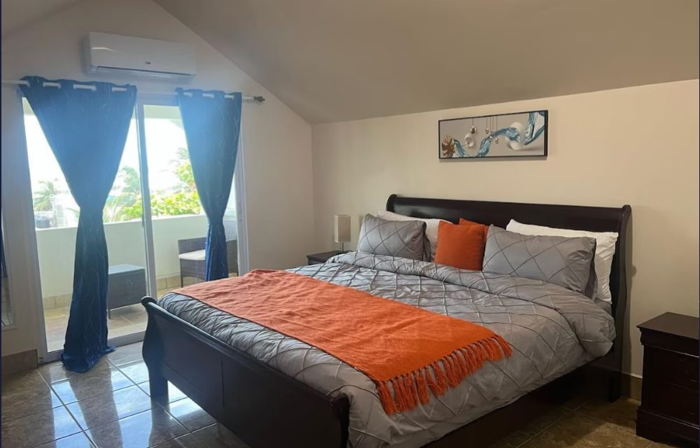 Deluxe Penthouse bedroom at Sea Horizon with a dark wood king bed, orange throw and balcony doors framed by blue curtains